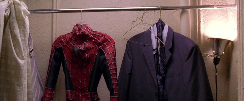 Cena do filme Homem-Aranha 2Na imagem, estão penduradas com cabides em uma barra de aço, o uniforme do Homem-Aranha e um terno com gravata, no meio dos dois há um cabide vazio. O uniforme está do lado esquerdo, ele é vermelho, mas possui detalhes em azul na parte inferior do braço e antebraço e também nas costelas. Ele possui uma aranha prateada no peito e teias em alto relevo por toda roupa. O terno é na cor azul. O armário é todo branco e tem aspecto de velho, no canto direito há uma luminária acesa apontada para cima