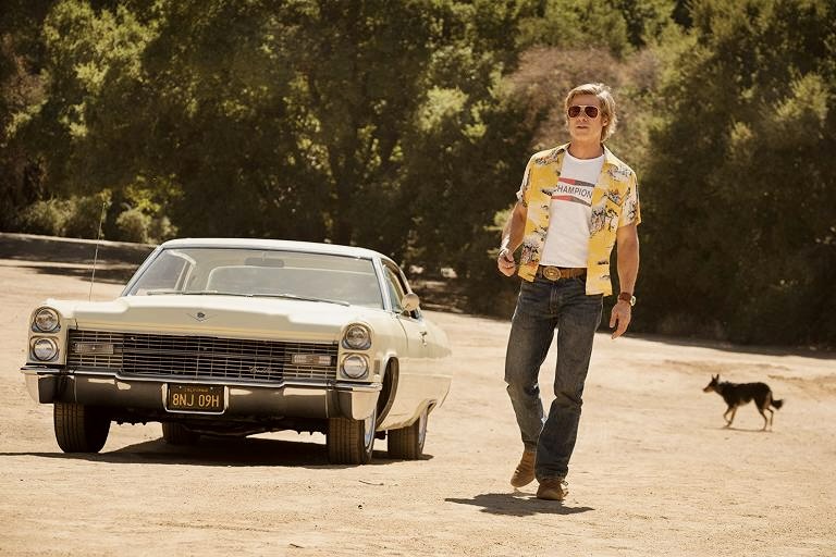 Cena de Era Uma Vez em… Hollywood. O personagem de Brad Pitt, homem branco e loiro que veste uma calça jeans, camiseta branca com detalhes vermelhos e uma camisa aberta amarela com detalhes florais e uma bota marrom, anda ao lado de um Cadillac creme, carro de 1966, com árvores ao fundo.