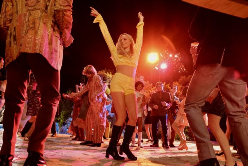 Cena de Era Uma Vez em… Hollywood. A atriz Margot Robbie, mulher branca e loira dança sorridente com os braços para cima, centralizada ao centro de uma pista de dança iluminada e rodeada de outras pessoas, vestindo um conjunto amarelo e botas pretas.