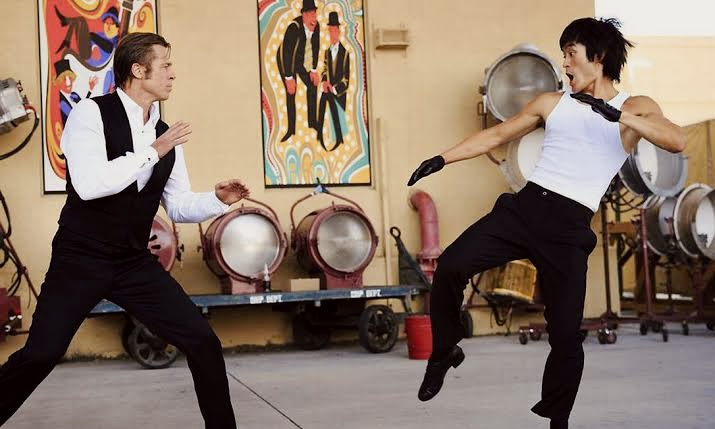 Cena de Era Uma Vez em… Hollywood. Bruce Lee (interpretado por Jason Scott Lee), homem de traços orientais vestindo uma regata branca e uma calça preta, faz movimentos de artes marciais enfrentando o personagem de Brad Pitt, homem branco que veste uma camisa branca de botões com uma calça preta e um colete preto.