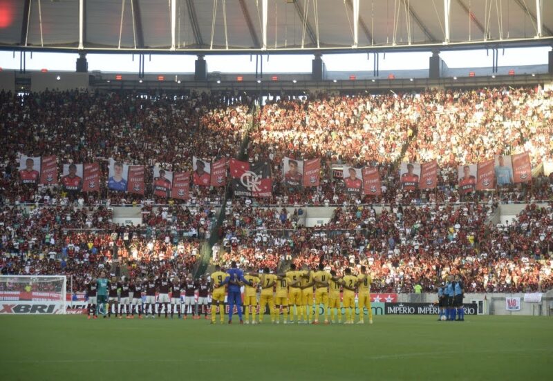 Jogo Flamengo contra Madureira no Maracanã em 2020, um ano após a tragédia. Em primeiro plano, vemos os jogadores abraçados e organizados em duas fileiras, uma de cada time. Ao fundo, a arquibancada está lotada de torcedores fazendo homenagem com cartazes para os meninos.