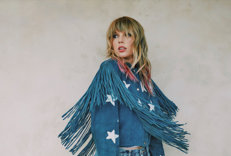 Taylor Swift posando contra uma parede clara, usando uma jaqueta azul com franjas longas e estrelas brancas estampadas. Seu cabelo está solto, com mechas rosadas nas pontas, e ela olha para o lado com uma expressão contemplativa.