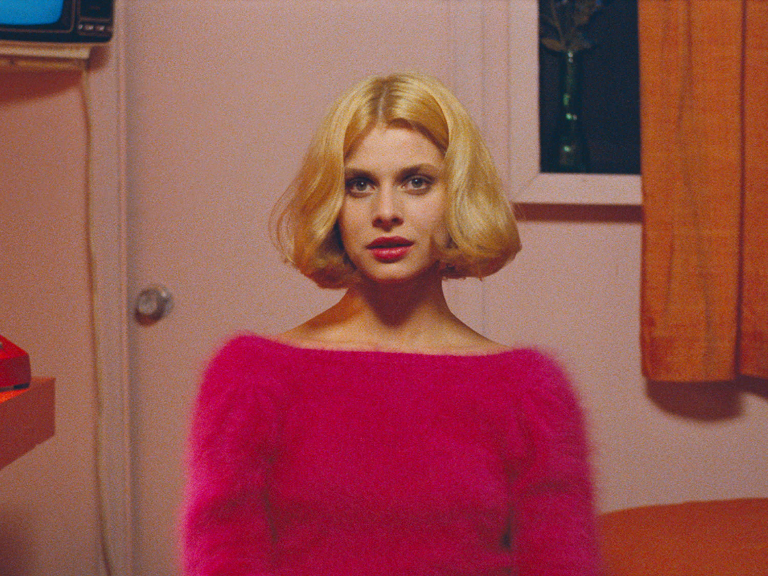 Imagem do filme Paris, Texas. Tem uma pessoa na imagem aparecendo da cintura para cima. No centro está Jane, uma mulher jovem e branca. Ela tem cabelo loiro na altura do pescoço, tem olhos castanhos e veste um suéter rosa felpudo. No fundo tem uma parede com uma porta e uma janela, o fundo também é rosa.