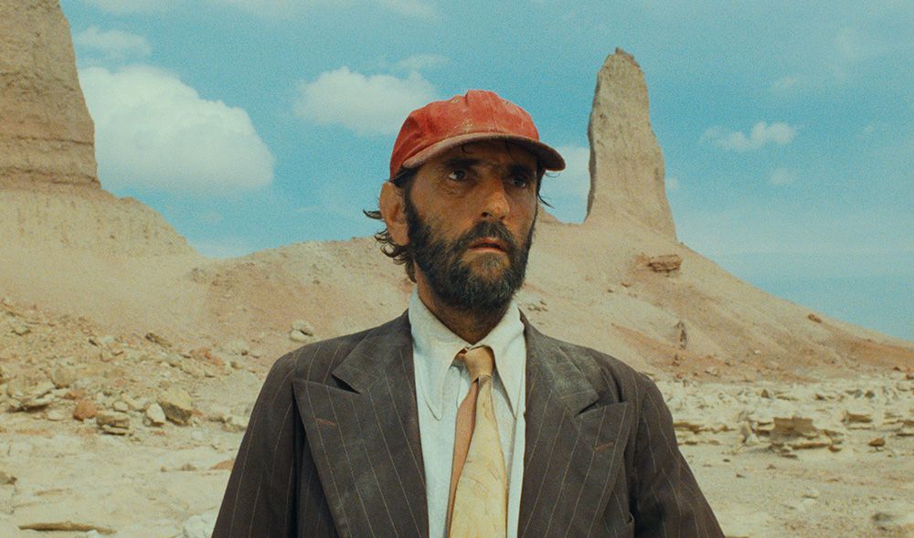 Imagem do Filme Paris, Texas. Tem uma pessoa no centro da imagem, aparecendo apenas o busto. No centro está Travis, um homem de meia idade. Ele tem cabelo curto e castanho, tem uma barba castanha e olhos castanhos. Ele usa um boné vermelho na cabeça e veste uma camisa branca, uma gravata amarela e um paletó cinza. Toda a sua roupa está suja de poeira. No fundo tem um deserto e o céu azul.