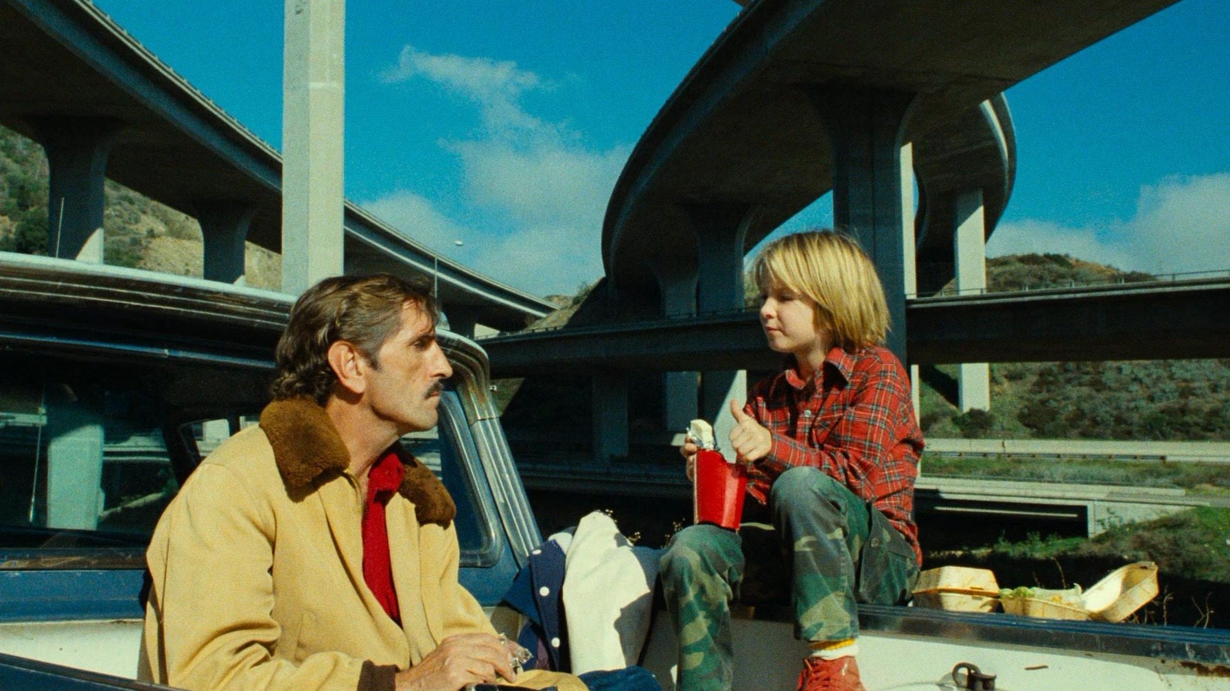 Imagem do filme Paris, Texas. Tem duas pessoas na imagem. Na esquerda, aparecendo o busto, está Travis, ele está com um bigode, ele usa uma jaqueta marrom e uma camisa vermelha. Na direita está Hunter, um menino de cabelo loiro comprido e de olhos castanhos, ele veste uma camisa xadrez vermelha, uma calça camuflada e um tênis vermelho. A criança tem um pote de batata frita apoiado na perna direita. Eles estão sentados na traseira de uma caminhonete conversando e se olhando. No fundo tem uma rodovia.Mostrar menos 