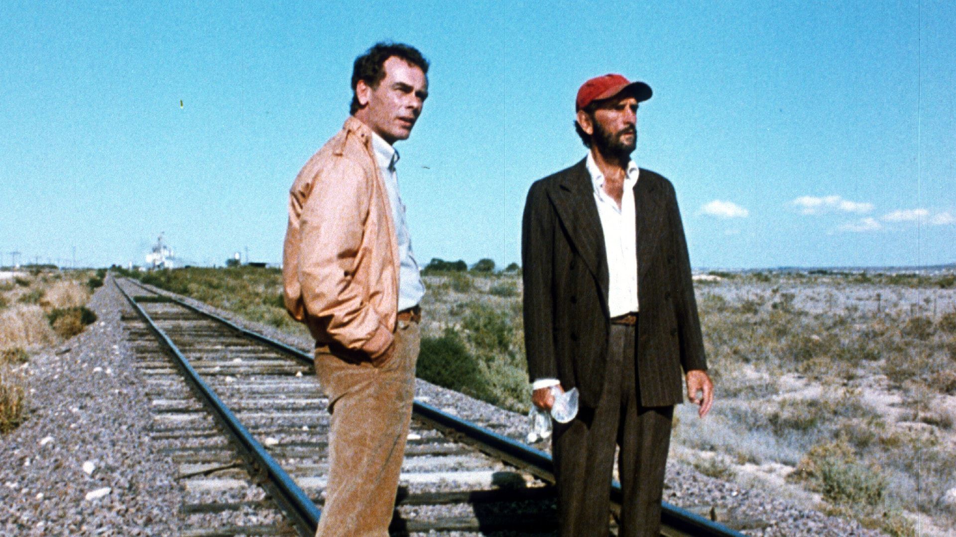 Imagem do filme Paris, Texas. Tem duas pessoas na imagem. Na esquerda está Walt, um homem de meia idade. Ele tem cabelo castanho curto e olhos castanhos, ele veste uma camisa azul, uma jaqueta marrom e uma calça marrom. Na direita está Travis, ele está de barba, ele veste um boné vermelho, uma camisa branca, um paletó cinza e uma calça cinza. Eles estão em cima de um trilho de trem e no fundo tem um deserto. Eles olham para o horizonte.