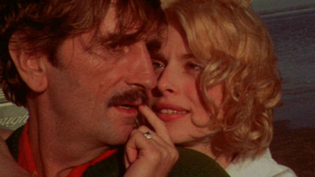 Imagem do filme Paris, Texas. Tem duas pessoas na imagem aparecendo somente o rosto. Na esquerda está Travis, ele veste uma camisa vermelha e uma jaqueta verde. Na direita está Jane, ela usa uma camisa branca. Jane abraça Travis por trás e está mexendo no bigode de Travis com a mão esquerda. No fundo tem um mar.