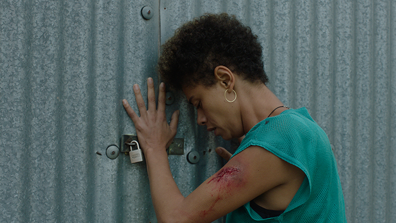 Cena do filme Continente. Na foto vemos Helô fechando um container. Helô, mulher negra de cabelos castanhos curtos, usa uma blusa azul bebê. Sua mão esquerda e o restante de seu corpo estão apoiados na porta de um grande container de metal.