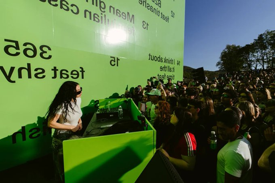 Fotografia de uma apresentação ao ar livre da cantora Charli Xcx, uma mulher  de cabelos pretos e blusa branca que está atrás de uma mesa de som verde, tocando para uma grande multidão. Ao fundo, uma estrutura verde-limão exibe textos pretos invertidos e desordenados. A luz do sol ilumina o ambiente, e várias pessoas na plateia parecem estar aproveitando o evento, usando óculos escuros e roupas casuais.