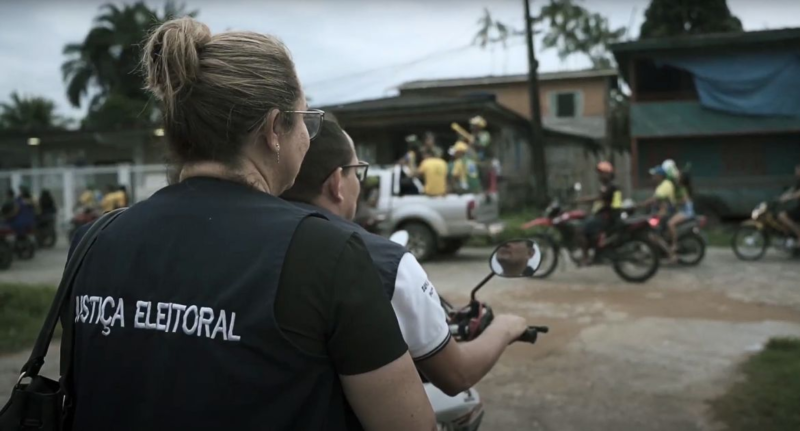 Cena do filme No Céu da Pátria Nesse InstanteNa imagem, um homem dirige uma moto, enquanto uma mulher com um colete escrito Justiça Eleitoral nas costas está na garupa. Eles estão de costas e na frente deles, em desfoque, há uma caminhonete com pessoas em cima que vestem camisetas verdes e amarelas, uniformes da seleção de futebol brasileira e usam bandeiras do país. Há também motos com pessoas que vestem o mesmo tipo de roupa. Também é possível ver casas no horizonte. A mulher e o homem estão na faixa dos 40 anos. 