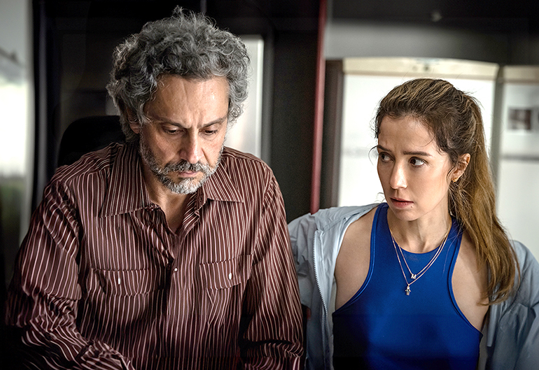 Cena do filme Precisamos Falar. Na imagem vemos Sandra e Paulo pensando em algo. Sandra, mulher branca com cabelos castanhos, veste um top azul e corta-vento cinza, seus cabelos estão presos em um rabo de cavalo. Celso, homem branco com barba e cabelos grisalhos, veste uma camisa de botão listrada branca e vinho. Ao fundo dos dois é possível ver uma geladeira.