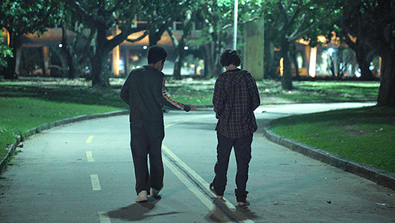 Cena do filme Precisamos Falar. Na imagem vemos Jefferson e Rick caminhando em um parque. Jefferson, adolescente negro com cabelo preto, usa blusa manga longa verde, calça preta e um tênis branco. Rick, adolescente branco de cabelos castanhos, veste camisa xadrez, calça e tênis ambos preto. Os dois estão caminhando de costas para a câmera.