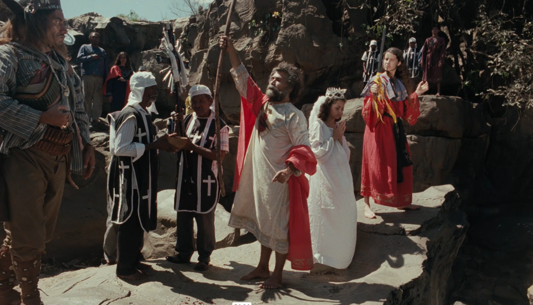 Na imagem, vemos uma cena do filme Corisco e Dadá onde ocorre um ritual relacionado ao pedido de batismo do filho do casal. Chico Diaz, como Corisco, observa com seriedade, vestindo trajes de cangaceiro. Ao centro, figuras com vestes litúrgicas conduzem a cerimônia, enquanto um homem de túnica clara ergue um cajado. Ao lado, uma mulher em oração e outra segurando uma espada reforçam a atmosfera solene. O cenário rochoso e árido do sertão contrasta com a espiritualidade do momento.