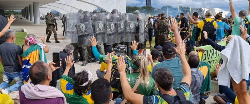 Cena do filme No Céu da Pátria Nesse InstanteNa imagem, muitas pessoas vestindo bandeira do Brasil e verde e amarelo estão em volta do exército brasileiro, que tenta impedir o avanço das pessoas. Elas estão filmando com o celular e levantando as mãos. O exército está em pouca quantidade e se protege com escudos.