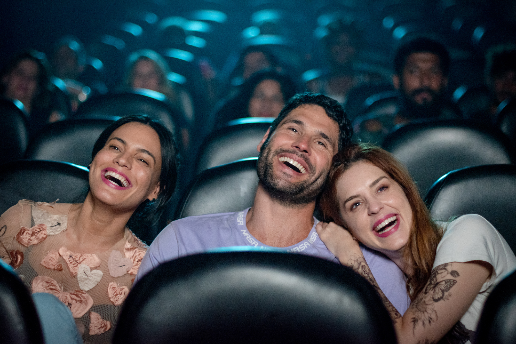 Cena do filme Todo Mundo (Ainda) Tem Problemas Sexuais. Em uma sala de cinema, três pessoas estão sentadas lado a lado, rindo de forma descontraída. À esquerda, uma mulher de cabelo escuro e vestido claro com bordados de corações sorri amplamente. No centro, um homem com barba curta e camisa lilás exibe um sorriso aberto, transmitindo alegria. À direita, uma mulher ruiva com tatuagens nos braços repousa a cabeça no ombro do homem, sorrindo de maneira suave e afetiva. Ao fundo, outras pessoas estão dispersas pelas poltronas, contribuindo para o clima leve e divertido da cena, evocando momentos de companheirismo e descontração compartilhada.