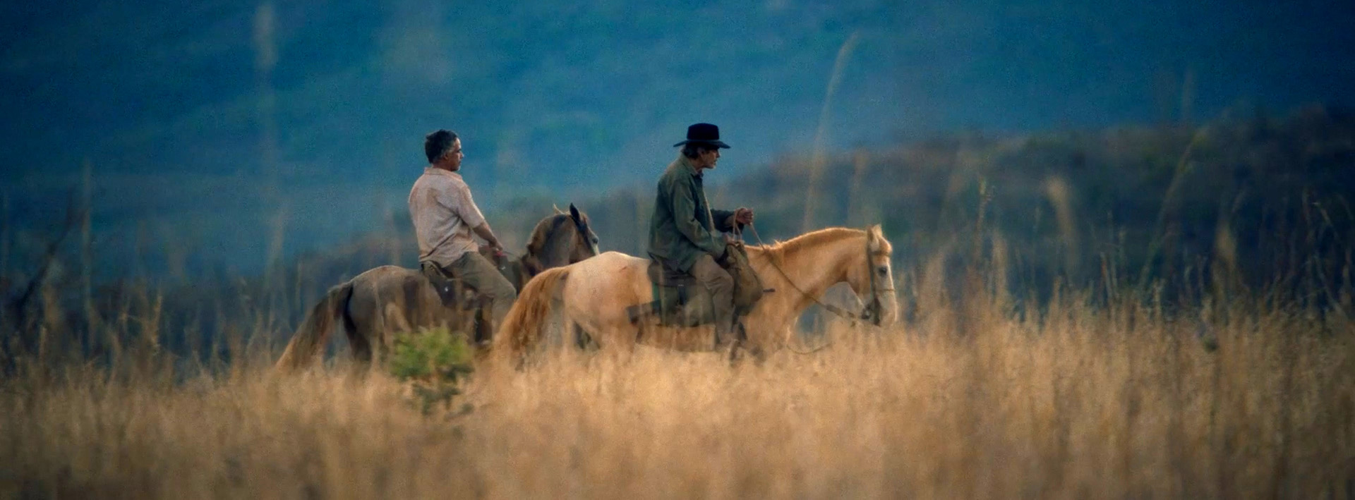 Na imagem, em formato horizontal, Toto e Jerominho cavalgam por uma paisagem de mato amarelado e morros ao fundo. O fundo é azul com árvores. Toto está do lado esquerdo, mais atrás de Jerominho, em cima de um cavalo com pelagem cinza. Ele veste uma camisa de botão na cor branca e calça jeans. Ele é um homem na faixa dos 60 anos, de pele escura e cabelos grisalhos. Jerominho está mais à frente, montado em um cavalo branco, bem sujo. Ele veste uma jaqueta azul e calça cinza, Usa na cabeça um chapéu preto. Ele é um homem na faixa dos 80 anos. 