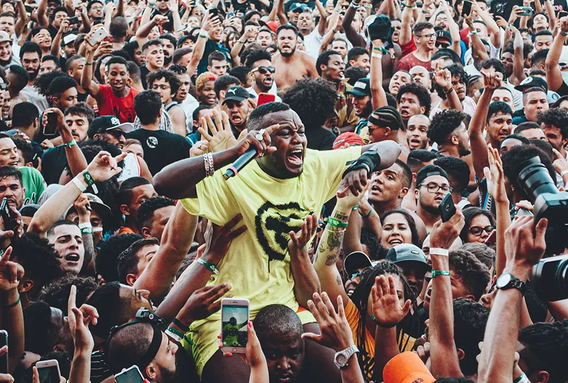 A imagem é uma fotografia do cantor Djonga. Djonga é um homem negro de cabelo crespo com a cor preta, e bigode, também preto. Na foto, o cantor está no meio da plateia de seu show, rodeado por centenas de pessoas e levantado por alguns dos presentes. Djonga veste uma camiseta e shorts na cor amarelo neon, e segura um microfone na mão direita.