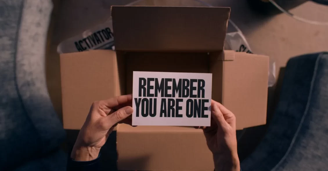 Cena do filme A Substância. Na imagem vemos as mãos de uma pessoa branca segurando um papel branco com a frase “REMEMBER YOU ARE ONE” escrita em preto. Atrás do papel encontra-se uma caixa de papelão com uma fita transparente escrita “ACTIVATOR” em preto. O carpete do chão está na cor cinza.