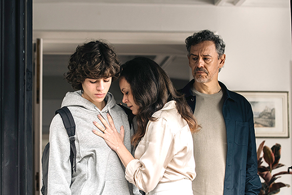 Cena do filme Precisamos Falar. Na foto vemos Rick, Anna e Celso se despedindo. Rick, adolescente branco com cabelos castanhos, veste um moletom cinza e em suas costas carrega uma mochila preta. Anna, mulher branca de cabelos castanhos, veste uma camisa clara. Sua mão esquerda segura o peito de seu filho Rick. Celso, homem branco com barba e cabelos grisalhos, veste uma camiseta cinza sobreposta por uma camisa de botões azul. Anna e Celso estão se despedindo de Rick. No fundo é possível ver uma parede branca com um quadro ao centro.