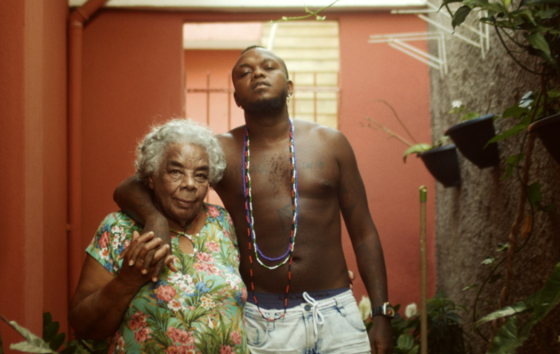 A imagem é uma peça promocional. Na foto, Djonga, um homem negro de cabelo crespo na cor preta, e cavanhaque, também preto, abraça sua avó, Maria Viana, uma mulher negra idosa, com cabelos na cor branca. Djonga está sem camiseta, veste um shorts jeans na cor branca, e usa três guias da umbanda, que são colares que se estendem até a altura do umbigo. Sua avó veste um vestido verde claro, estampado com diversas flores rosas e brancas, além de alguns ramos verdes. Aos fundos, há uma parede marrom, além de alguns vasos pendurados, no canto direito da imagem.