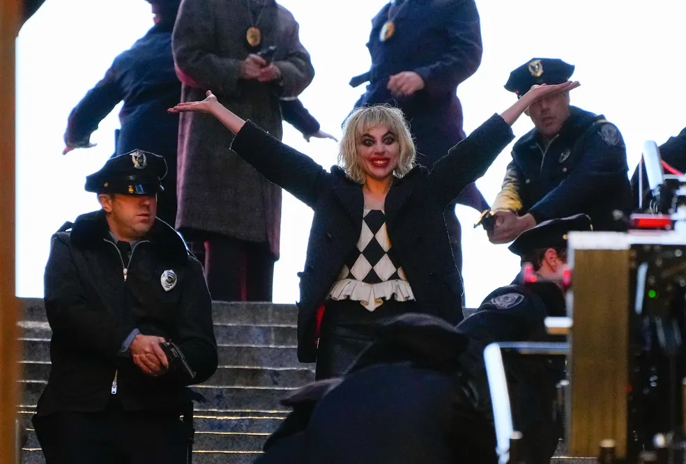 Foto da cantora Lady Gaga atuando no set de Coringa: Delírio a Dois como Arlequina.  Ela está usando uma blusa preta e branca em padrão de losangos, típica do figurino clássico da personagem, e um casaco escuro por cima. Seu cabelo é loiro platinado, com um corte curto e bagunçado, reforçando o estilo icônico da Arlequina. Ela está com uma expressão de euforia, braços abertos em um gesto amplo. Ao redor dela, há vários policiais uniformizados. A cena está ocorrendo em um ambiente público, em uma escadaria.