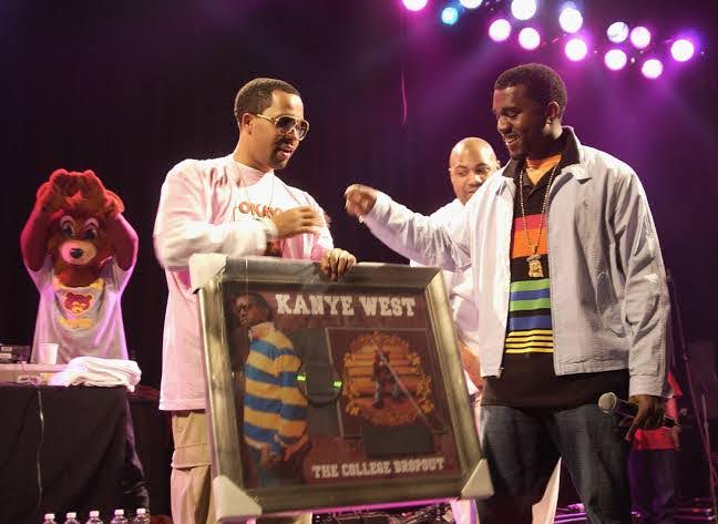 Foto do cantor Kanye West recebendo seu primeiro disco de Platina. A imagem é retangular com fundo preto. Na parte esquerda, há uma pessoa vestindo uma fantasia felpuda de urso marrom com olhos marrons e focinho bege, camiseta cinza estampada com a cabeça de um urso marrom de pelúcia, calça preta e está com o braço levantado, fazendo com as mãos o símbolo de um coração. Logo à frente do urso, há uma mesa coberta por um pano de mesa preto e, em cima dela, um copo branco e algumas camisas brancas dobradas. Na parte central esquerda, um homem negro de cabelo raspado e cavanhaque, usando um óculos modelo aviador, vestindo uma blusa de manga comprida branca e calça cargo bege, está segurando um quadro emoldurado de cor dourada, com a foto do artista Kanye West em pose de triunfo, vestindo uma camisa de manga comprida com listras azuis e amarelas e, ao lado, as palavras “Kanye West The College Dropout” e a capa do álbum, composta por um mascote de urso marrom, em tamanho humano, sentado em madeiras marrom. Está vestindo um blazer marrom, moletom laranja e calças jeans. Suas mãos estão apoiadas no joelho. Enquadrando a foto, há uma moldura dourada com dois anjos em cada lado, encontrados na parte central dos lados. Ao lado do homem, na lateral direita da imagem, com o braço esticado para pegar a moldura, está Kanye West sorrindo. O artista tem um cavanhaque, está usando uma blusa preta com listras coloridas, jaqueta cinza e calças jeans e uma corrente de ouro.