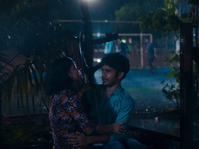 Cena do filme Tudo Que Imaginamos Como Luz. No fundo, uma quadra com vários garotos jogando futebol. O ambiente é escuro, com uma iluminação baixa. Mais próximo a câmera, como se fosse em um outro plano, estão Anu e Shiaz abraçados em frente a uma árvore. Anu veste uma roupa florida, com tons mais escuros. Shiaz veste uma roupa com tons azulados e detalhes em forma de árvore.