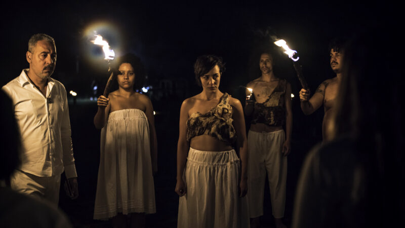 Cena do filme Corpo Presente. Na imagem, um grupo de cinco pessoas aparece enfileirado. No centro tem três mulheres e dois homens nas extremidades. As pessoas vestem roupas manufaturadas nas cores marrom e off white. Tudo é bem natural e duas das mulheres seguram tochas nas mãos. 