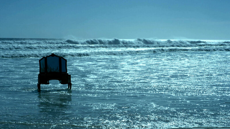 Cena do filme Corpo Presente. Na imagem, uma cadeira aparece abandonada na areia da praia, em um lugar que recebe as ondas da praia. O céu e o mar têm um tom de azul cristalino.