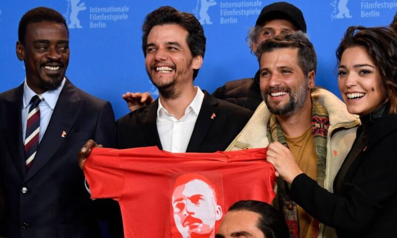 [Texto alternativo: Na foto, há quatro pessoas enumeradas da esquerda para a direita, sendo elas Seu Jorge, Wagner Moura, Bruno Gagliasso e Bella Camero, segurando uma camiseta vermelha de tom vibrante com o rosto de Carlos Marighella. O fundo atrás é de cor azul e é do Festival Internacional de Cinema de Berlim.]