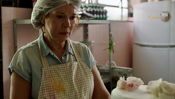 Cena do filme A procura de Martina. Na imagem está a mãe adotiva do neto de Martina. Ela é uma mulher com mais de cinquenta anos, de pele branca e marcas de expressão no rosto. Ela está com uma touca e um avental de cozinheira em frente a uma parede cor de rosa pálido.