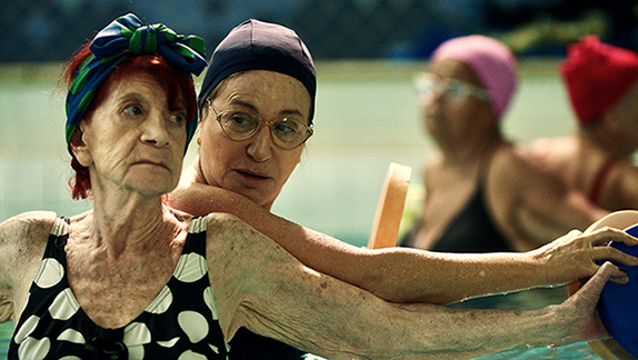 Cena do filme A Procura de Martina. Na imagem estão Matina e sua amiga. As duas são mulheres com mais de 60 anos, brancas e com cabelos castanhos. Elas estão na piscina com toucas de natação fazendo hidroginástica. 