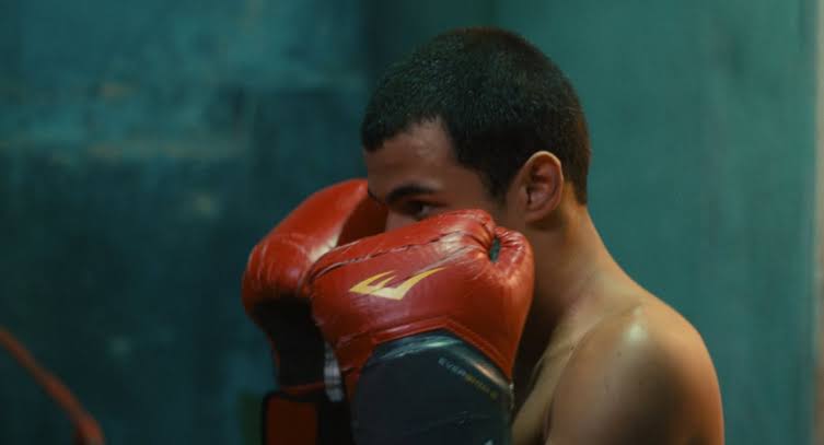 Cena do filme Baby Na imagem, o personagem Baby faz pose de guarda na luta de Boxe, colocando as duas mãos ao lado do rosto. As mãos estão vestidas com luvas de Boxe na cor vermelha. Baby é um garoto na faixa dos 18 anos, de pele escura e cabelos escuros, bem curtos. Ele está sem camiseta. Ao fundo há uma parede azul. 