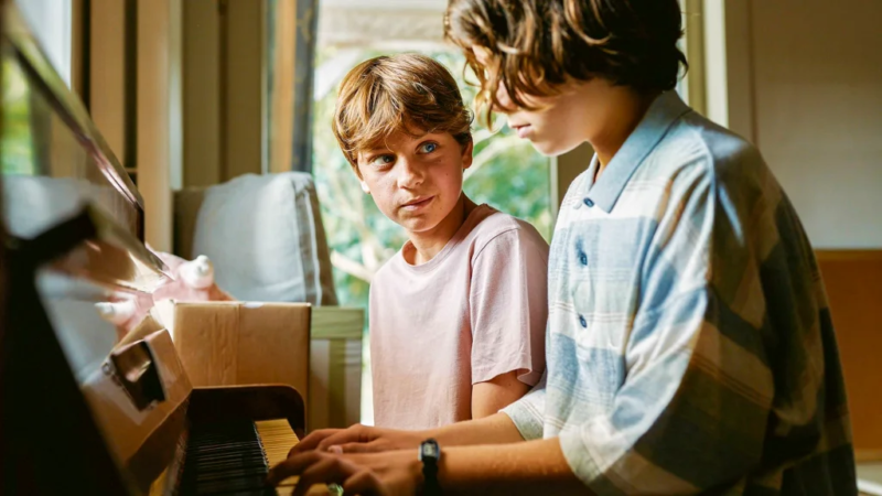 Cena do filme Corações Jovens. Lou Goossens e Marius de Saeger estão juntos ao piano, em uma cena que exala cumplicidade e curiosidade. Lou observa Marius com atenção enquanto ele toca, demonstrando interesse e admiração. A luz natural que entra pela janela ilumina o ambiente, criando uma atmosfera leve e serena, que destaca o vínculo e a troca de olhares entre os jovens.