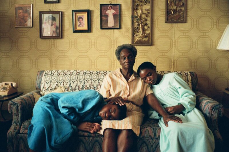 Cena do filme Todas as Estradas de Terra Têm Gosto de Sal. Três gerações de mulheres negras estão reunidas em um sofá em uma sala decorada com fotos de família na parede ao fundo. No centro, uma senhora de cabelos grisalhos e expressão serena segura com carinho as cabeças de duas meninas mais jovens, suas netas, uma de cada lado. À sua esquerda, uma garota de aproximadamente 10 anos, usando um roupão azul, descansa a cabeça em seu colo com os olhos fechados. À direita, outra jovem, com cerca de 12 anos e vestindo um roupão verde claro, também repousa a cabeça na perna da idosa, criando uma cena de aconchego, amor e conexão entre as gerações.