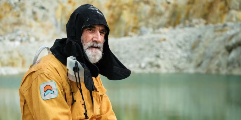 Cena de O Planeta Silencioso. Nela. Vemos Theodore, um homem branco de barba branca. Ele veste um macacão amarelo e uma touca preta. Ele esta olhando para o lado e ao fundo há uma pedreira e um lago