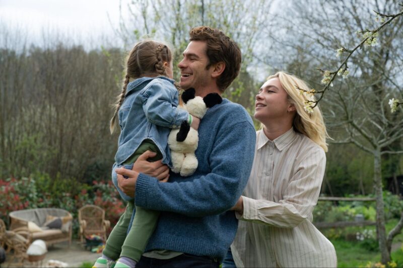 Cena do filme Todo Tempo Que Temos. Na imagem, Tobias, Almut e a filha do casal estão juntos em um jardim. Eles estão enfileirados, a criança está no colo de Tobias, olhando para trás, o rosto dela não está visível. Atrás de Tobias, na direita, está Almut. Eles estão felizes, sorrindo. Ela é uma mulher na faixa dos 30 anos, de pele clara e cabelos loiros, lisos e compridos. Ela está vestindo uma camisa social de botões na cor branca com listras pretas na vertical. Tobias é um homem branco, na faixa dos 40 anos, de cabelos curtos na cor castanho claro. Ele usa um suéter azul marinho. A criança também possui pele clara, cabelos castanhos com um penteado de tranças nos dois lados da cabeça. Ela usa uma jaqueta jeans e calça moletom na cor verde escuro. Na mão direita ela segura um ursinho de pelúcia com o corpo branco e orelhas e rabo na cor preta. O jardim possui muitas árvores secas, grama verde e flores vermelhas. No canto esquerdo há um espaço para descanso, com sofá com almofadas e uma cadeira de madeira.