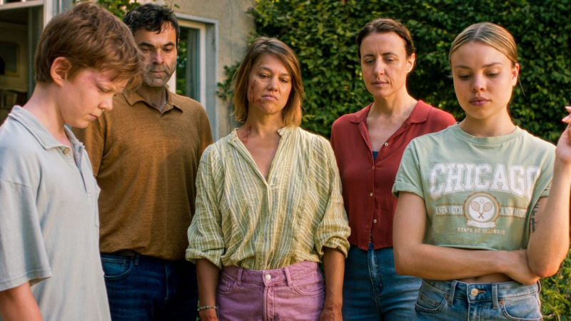 Cena do filme O Pardal na Chaminé. Cinco pessoas estão lado a lado, com expressões introspectivas e semblantes melancólicos, sugerindo um momento de reflexão ou tristeza. À esquerda, um jovem de cabeça baixa veste uma camisa clara, enquanto ao seu lado está um homem de barba curta, usando um suéter marrom. No centro, uma mulher de cabelos curtos e roupa leve, com uma blusa listrada e shorts roxos, olha para baixo com ar desanimado e rosto sujo de sangue. Ao lado dela, uma mulher de cabelo preso veste uma camisa vermelha, enquanto a última personagem, à direita, usa uma camiseta verde com a palavra "Chicago". A composição evoca uma atmosfera de tensão emocional, capturando um instante de silêncio e introspecção compartilhada pelo grupo. Ao fundo, há vegetação densa e uma parede clara, próximo a uma casa.