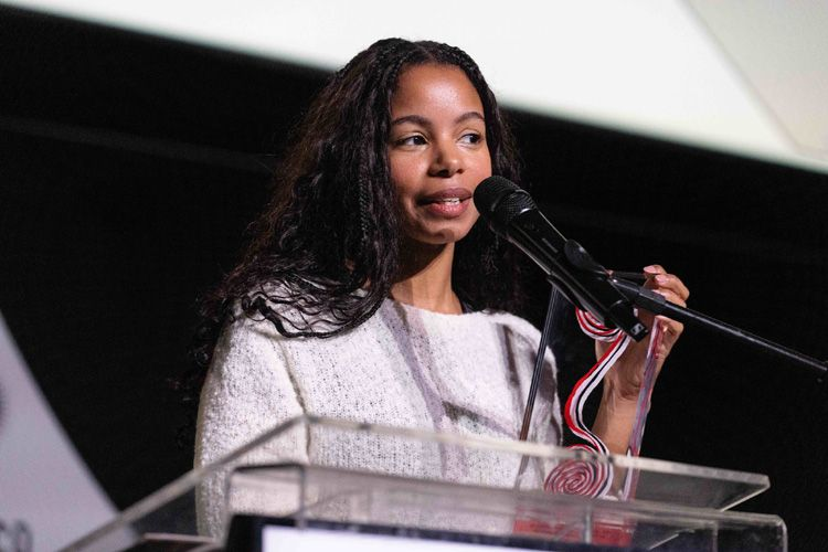 Denise Fernandes, diretora do filme Hanami, fala ao microfone no palanque da 48ª Mostra Internacional de Cinema de São Paulo. Ela é uma mulher negra de cabelos cacheados escuros e longos, vestindo um suéter claro com textura. Denise segura um troféu ao falar ao microfone, com um sorriso leve e uma expressão de concentração. O palco tem um fundo escuro, destacando a oradora.