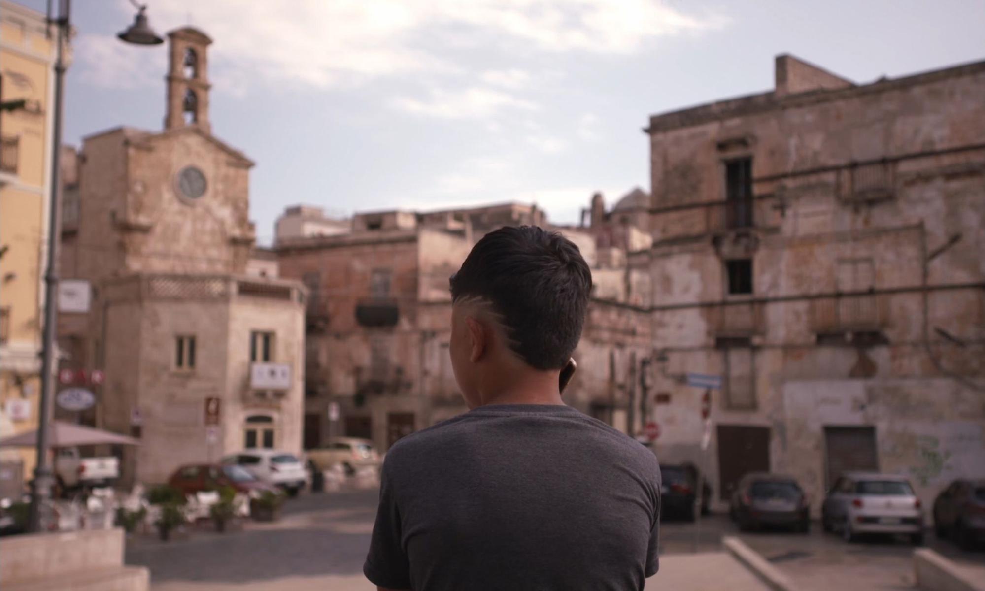 Cena do filme Bangarang. Na imagem, um menino está de costas para a câmera, observando a capital homônima da província de Taranto, localizada na Itália. A paisagem é formada por construções antigas e desbotadas, além de estar com uma cortina de poluição. A câmera captura o menino de costas a partir do busto, ele veste uma camiseta cinza. O cenário reflete abandono e descaso das autoridades.