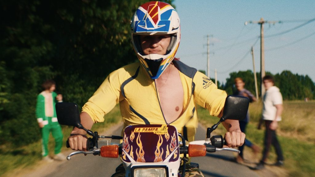 Cena do filme La Pampa. Amaury Foucher, de cabelo loiro e vestindo uma jaqueta amarela com detalhes pretos, está pilotando uma moto com um capacete colorido. Ele tem uma expressão séria enquanto segue pela estrada. No tanque da moto, vê-se o nome "La Pampa" e uma pintura de chamas. Ao fundo, algumas figuras desfocadas observam ao longe, enquanto a estrada e os postes de energia completam a ambientação rural, e seus amigos o olhando em alta velocidade.