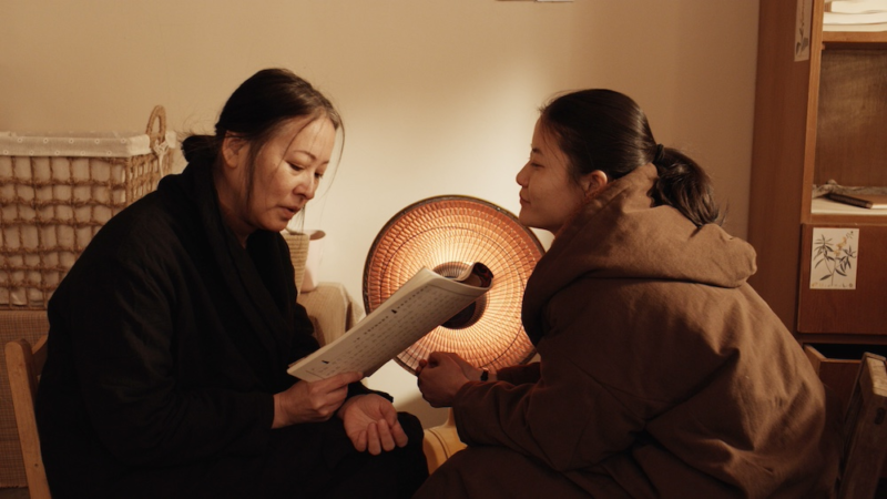 Cena do filme Uma Terra Deixada para Trás. Em um ambiente interno acolhedor e levemente iluminado, duas mulheres estão sentadas próximas, engajadas em uma conversa íntima. A mulher à esquerda, com cabelo preso e vestindo um casaco escuro, segura um papel e observa atentamente enquanto fala. A mulher à direita, usando um casaco marrom, inclina-se em direção à outra com uma expressão atenta e ligeiramente sorridente. Ao fundo, uma luminária de luz quente cria um ambiente aconchegante, destacando os detalhes das expressões e sugerindo um momento de conexão e compreensão mútua entre as duas personagens.