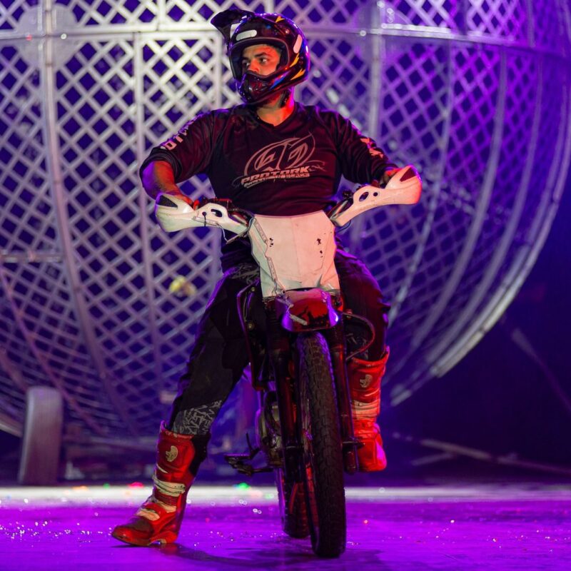 A imagem retrata um dos motociclistas se preparando para o número do Globo da Morte. Ele está equipado com capacete e roupas de proteção, com a estrutura metálica do globo ao fundo. A expressão focada e a postura do piloto refletem a concentração necessária para a execução deste perigoso número, um dos mais esperados do espetáculo.