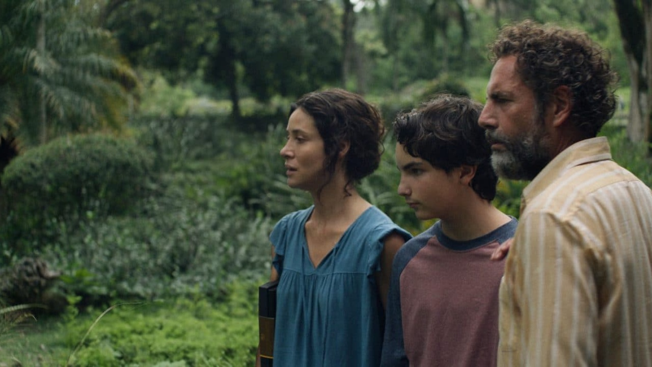 Cena do filme Zafari. Na cena vemos Ana, Bruno e Francisco olhando para algo. Ana, mulher branca com cabelos castanhos, veste uma blusa na cor azul. Bruno, adolescente branco de cabelos castanhos, usa uma camiseta de manga longa vermelha e cinza. Francisco, homem branco com cabelos e barba grisalhos, usa uma camiseta de botão listrada branca e amarela. Todos encontram-se na lateral direita da imagem, e, ao fundo, é possível ver árvores e vegetação.