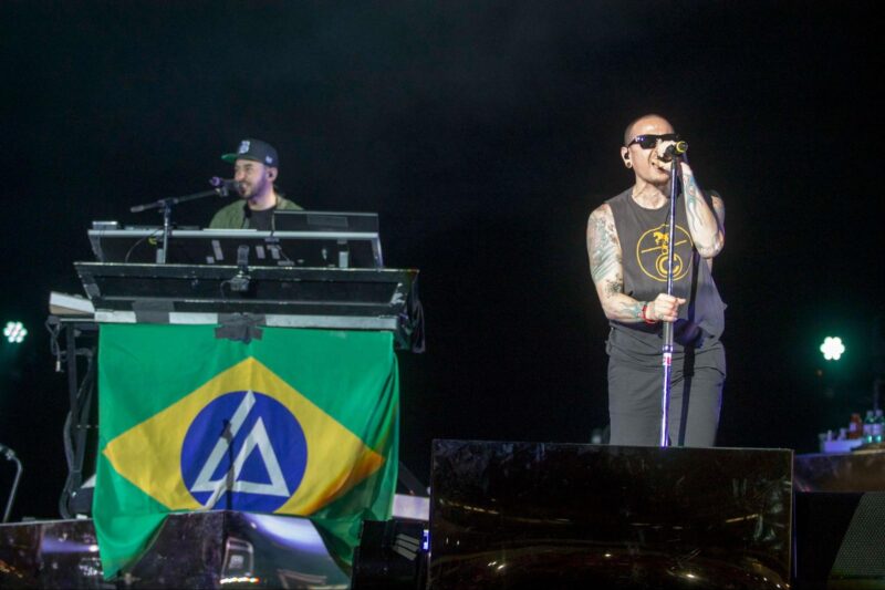 Foto de show do Linkin Park no Brasil. Na imagem, aparecem Chester e Mike em cima do palco, e ambos estão cantando. Em foco, do lado direito da imagem está Chester, que segura um microfone de chão com as duas mãos. Ele é um homem branco, careca e com tatuagens nos dois braços. Veste uma calça preta, uma regata preta com um círculo estampado no meio e alguns detalhes dentre dele e um óculos escuro quadrado. Ao fundo, do lado esquerdo da imagem e um pouco desfocado, está Mike, atrás de um apoio com um teclado em cima. Ele é um homem branco com ascendência japonesa e tem barba castanha. Ele veste uma camiseta verde musgo e um boné preto com detalhes em verde. Na frente de Chester está uma caixa de som grande e metalizada, que cobre ele até os joelhos, e junto com o teclado na frente de Mike está uma bandeira do Brasil com o logotipo do Linkin Park dentro da parte azul da bandeira. O fundo é escuro e bem nos cantos centrais tem dois iluminadores de palco verdes.