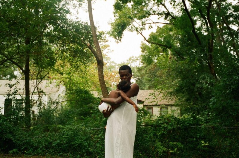 Cena do filme Todas as Estradas de Terra Têm Gosto de Sal. Uma mulher negra, vestindo um vestido branco, está em pé em um campo verdejante, segurando sua filha nos braços. Ela está envolta em um pano branco e deitada confortavelmente no colo da mulher, que olha para ela com ternura e proteção. A figura da mulher transmite força e suavidade, enquanto o fundo de árvores e casas distantes cria uma atmosfera serena e natural. A luz suave destaca o laço afetivo entre elas, capturando um momento de amor e cuidado materno.