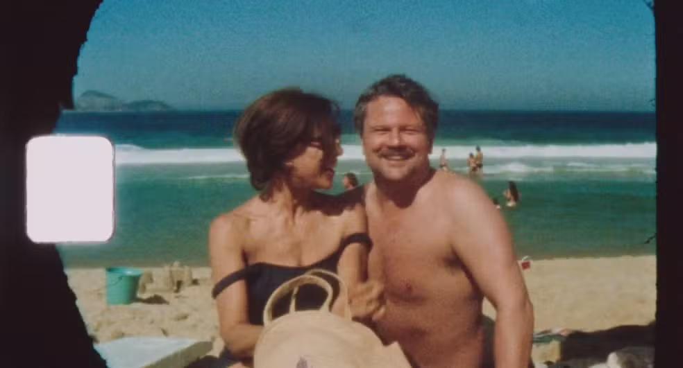 Na imagem, Eunice Paiva, mulher branca de cabelo curto castanho, olha para Rubens Paiva, homem branco de cabelo curto e com bigode, enquanto ambos posam para uma fotografia na praia. Ela veste um conjunto preto e está sorrindo para o homem. Ele está sem camiseta e também sorri em direção à câmera. 