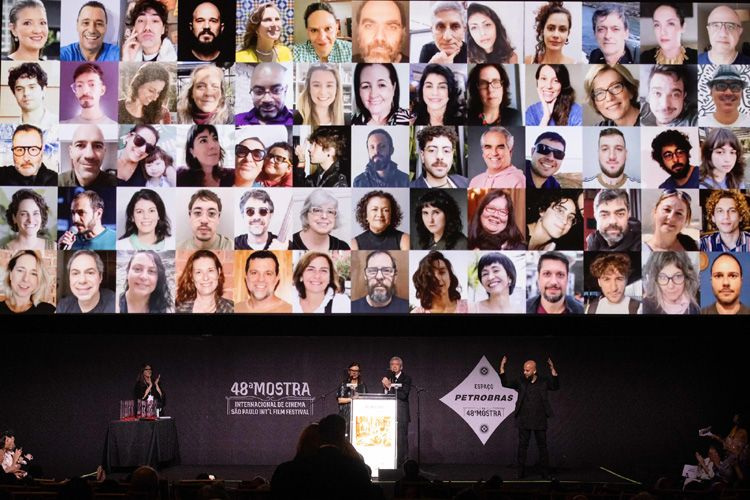 Painel com fotos de diversos rostos da equipe projetados em um telão durante a cerimônia da 48ª Mostra Internacional de Cinema de São Paulo. Abaixo, Serginho Groisman e Renata Amaral