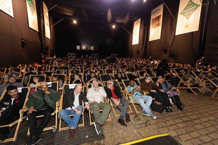 A imagem mostra o espaço Petrobras na Cinemateca Brasileira, uma grande sala de cinema com fileiras de cadeiras de madeira estilo espreguiçadeiras, onde o público está sentado, aguardando o início da sessão. No primeiro plano, várias pessoas estão sentadas, algumas em conversa e outras olhando para frente. À esquerda, um homem de jaqueta verde e chapéu está relaxado em sua cadeira, enquanto outros ao lado interagem entre si. O público ao fundo ocupa quase todas as cadeiras disponíveis. As paredes são pretas, e banners com logotipos e informações do evento estão pendurados nas laterais. O espaço é iluminado por luzes focais no teto, criando um ambiente adequado para a exibição de filmes.