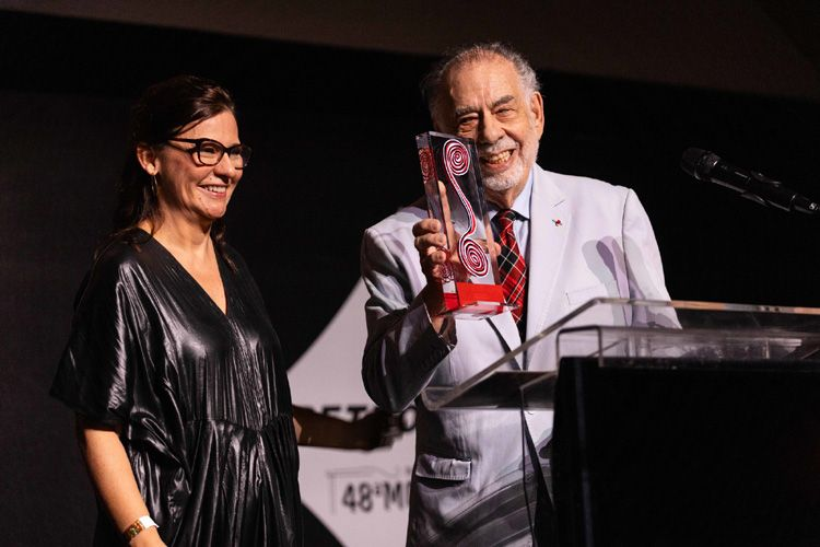 Francis Ford Coppola, sorrindo, segura um troféu em forma de espiral ao lado de Renata Almeida, diretora da Mostra. Coppola, um homem mais velho, com cabelo grisalho e barba branca, veste um terno branco com camisa e gravata vermelha com padrão xadrez. Ao seu lado, Renata Almeida, uma mulher de cabelos castanhos escuros presos, usa óculos de armação fina e veste um vestido escuro com aparência metálica. Ela sorri para Coppola enquanto segura o troféu. Ambos estão em roupas formais durante a cerimônia de premiação da 48ª Mostra Internacional de Cinema de São Paulo, com um banner do evento ao fundo.